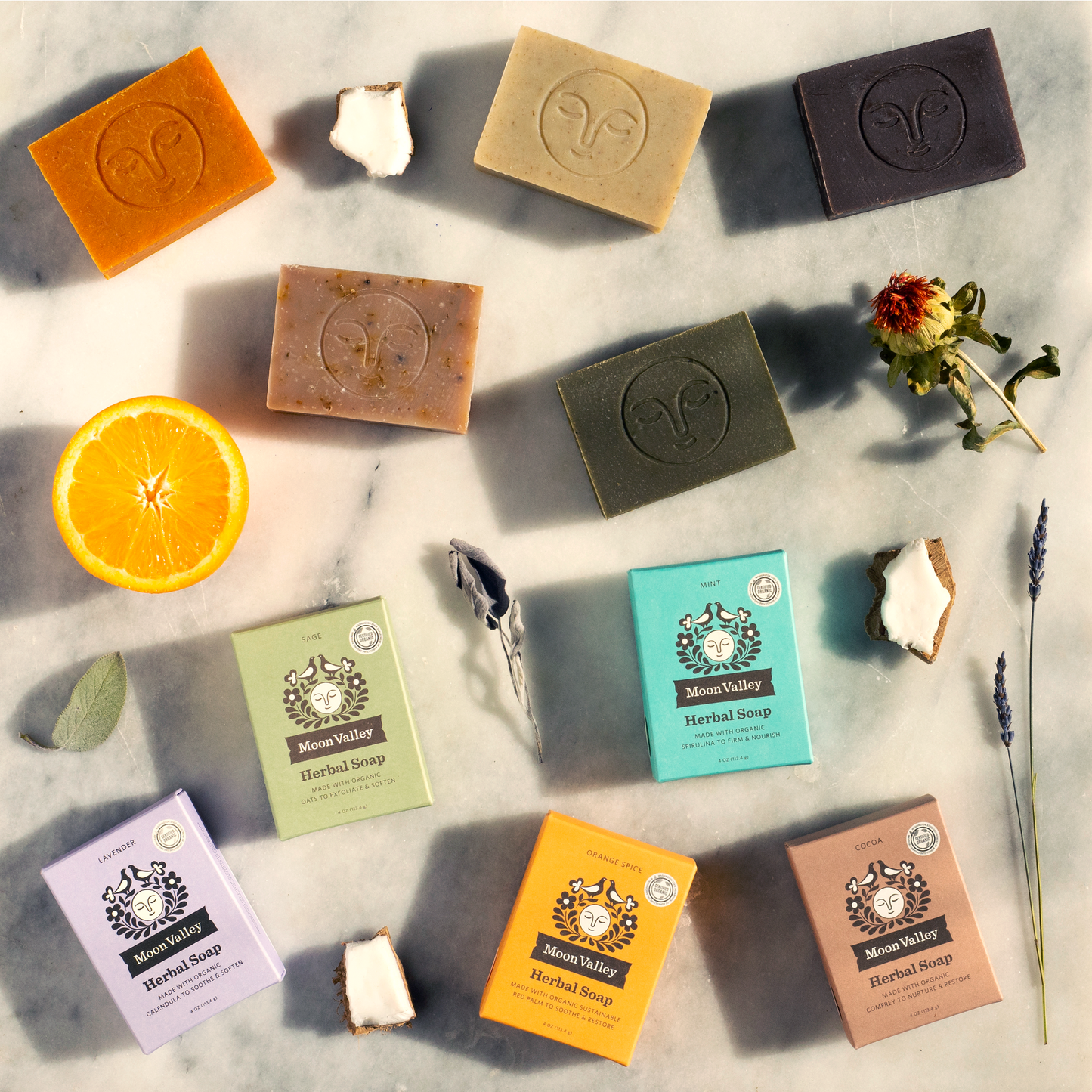 Moon Valley Organics Herbal Soap Bar Assortment of all bar soaps and their boxes on a counter with pieces of orange and sage and lavender and mint and coconut and safflower