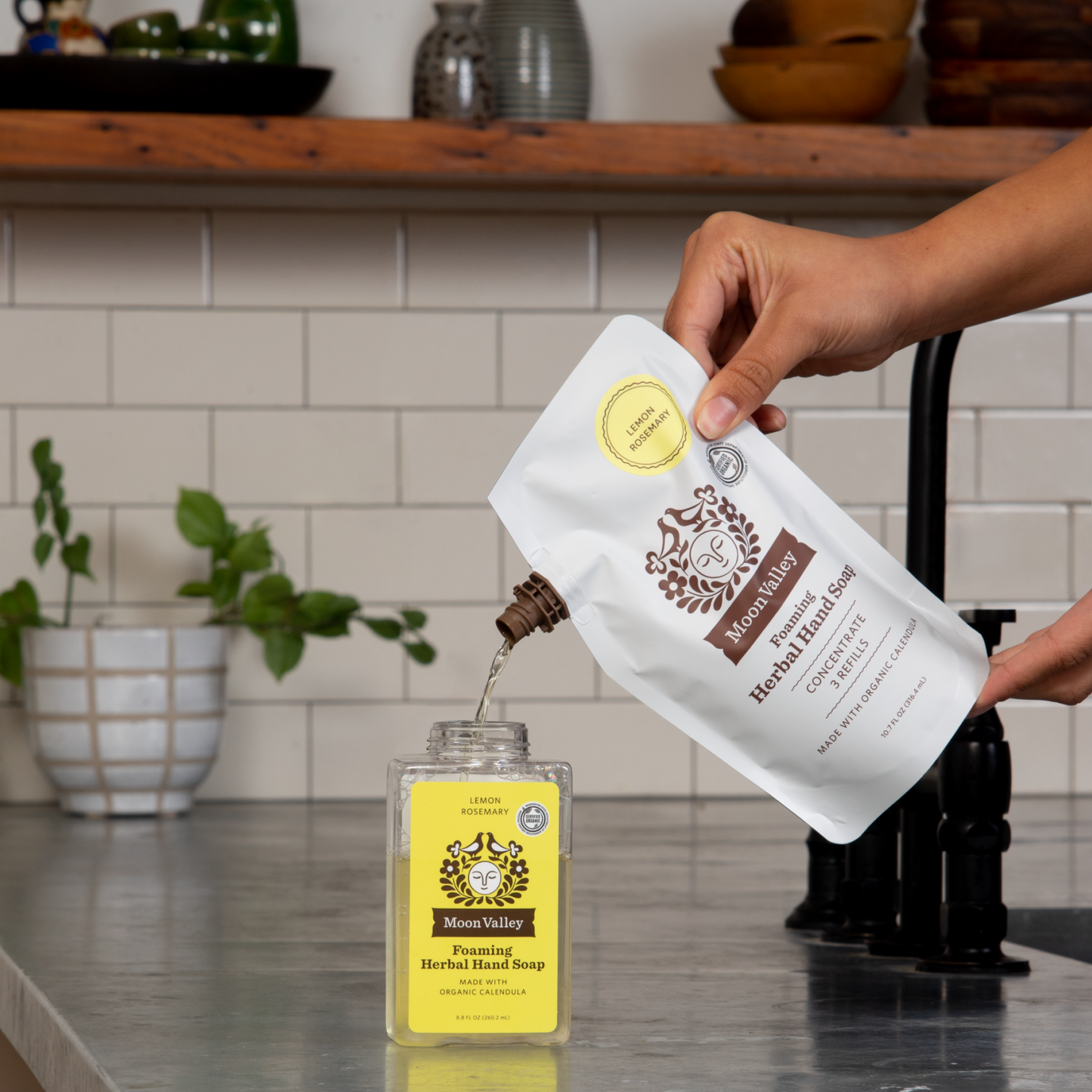 Moon Valley Organics Foaming Herbal Hand Soap Refill being poured into Foaming Herbal Hand Soap Dispenser Lemon Rosemary on a kitchen sink