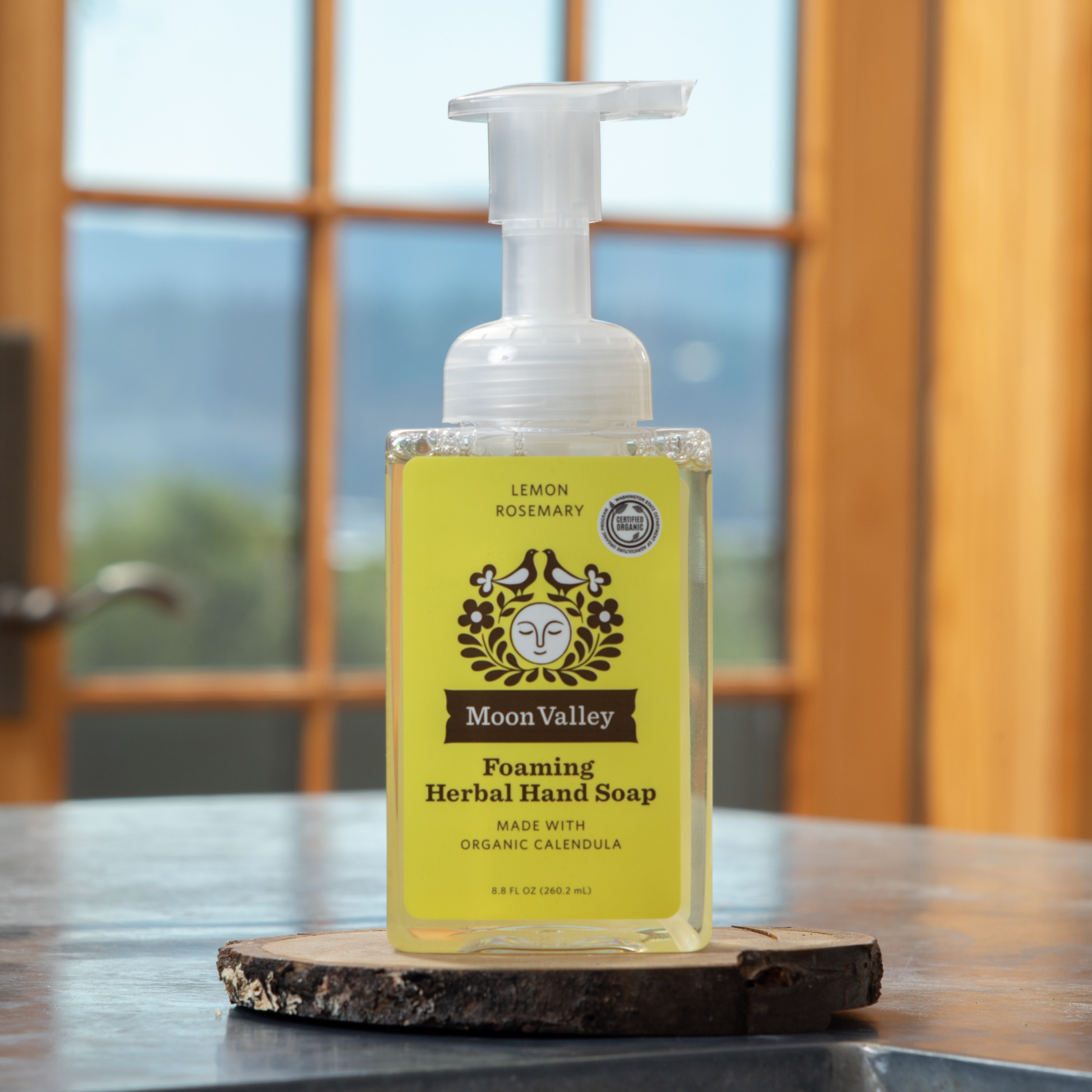 Moon Valley Organics Foaming Herbal Hand Soap Front Dispenser Bottle Lemon Rosemary sitting on a wooden coaster on a kitchen sink