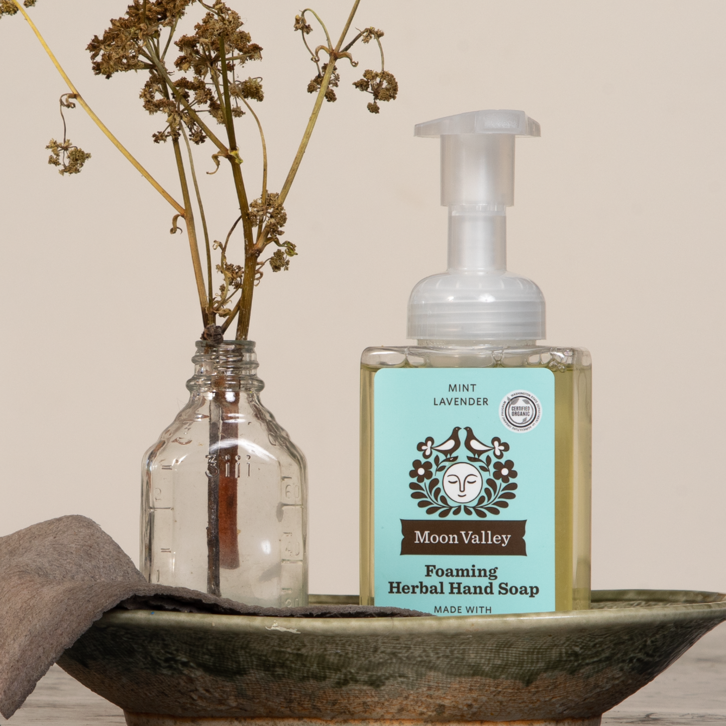 Moon Valley Organics Foaming Herbal Hand Soap Front Dispenser Bottle Mint Lavender with a bottle of dried flowers and a towel in a dish
