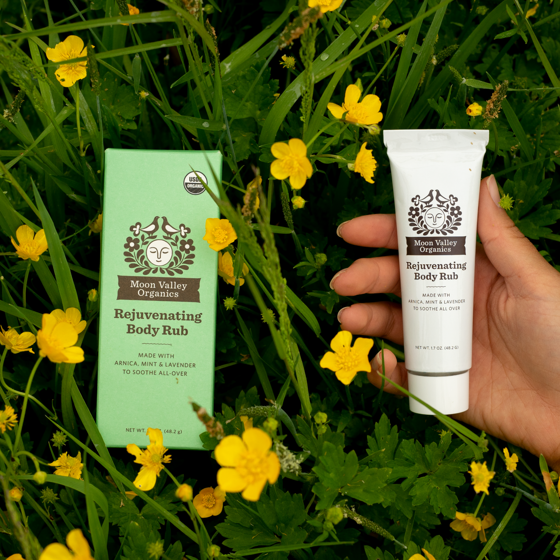 Moon Valley Organics Rejuvenating Body Rub Box laying in the grass and flowers while a hand holds the tube