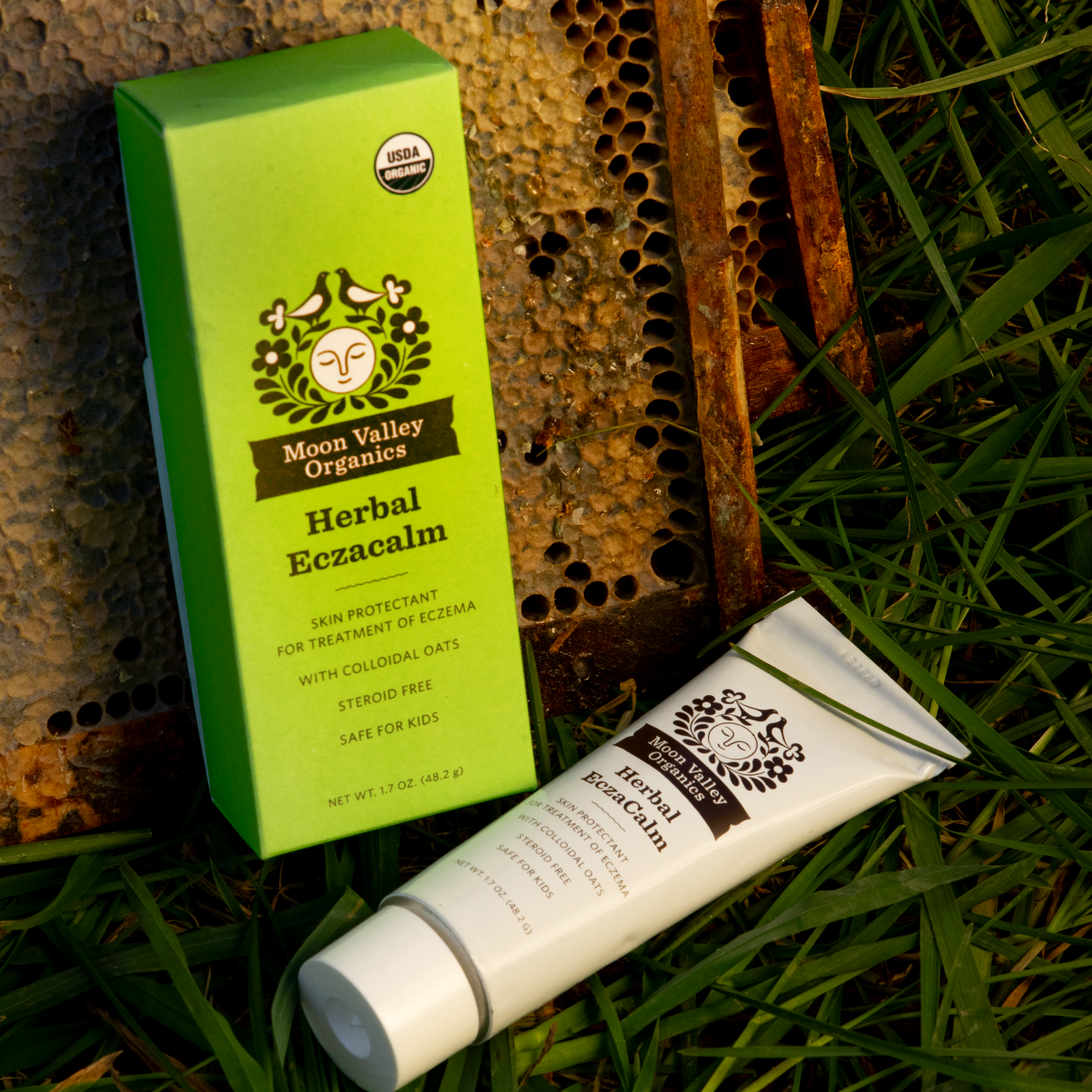 Moon Valley Organics Herbal Eczacalm Box and tube laying in the grass against a bee box and honeycomb