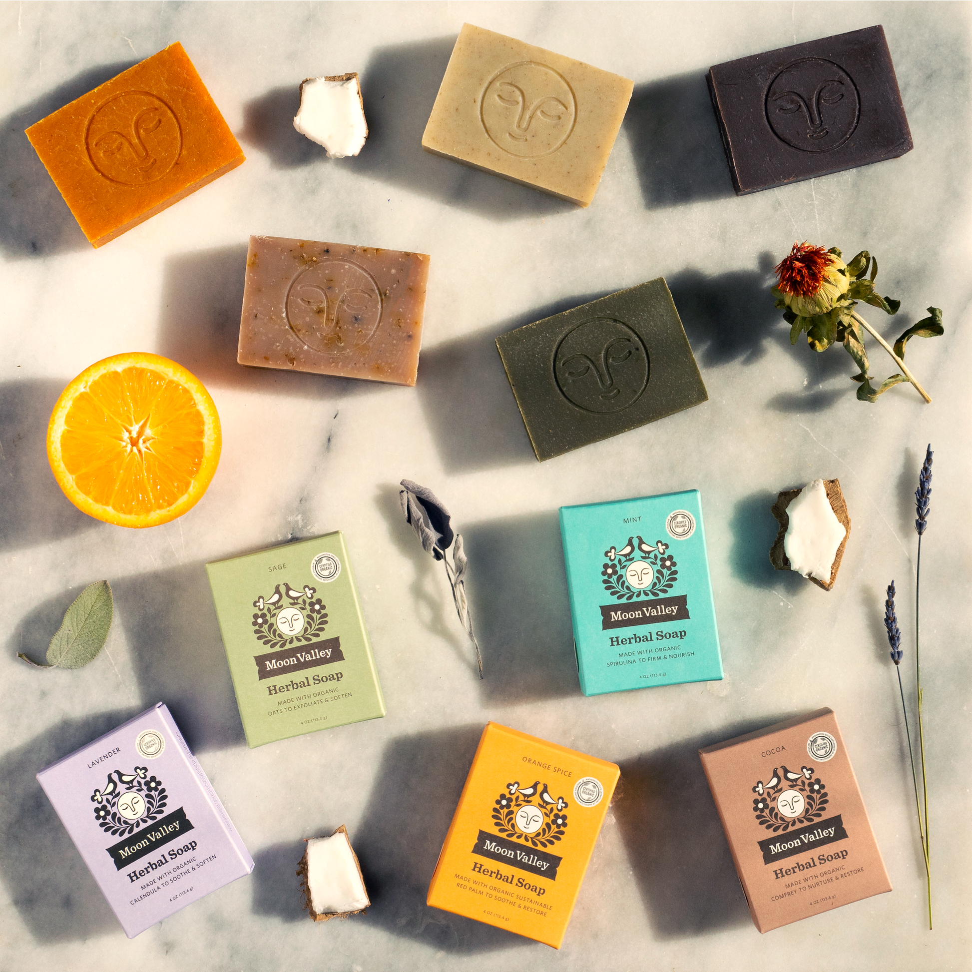 https://www.moonvalleyorganics.com/cdn/shop/files/4MoonValleyOrganicsHerbalSoapBarAssortmentofallbarsoapsandtheirboxesonacounterwithpiecesoforangeandsageandlavenderandmintandcoconutandsafflower_32f35c90-f130-4dc7-a3f8-39468ab442bc.png?v=1696952099&width=1946