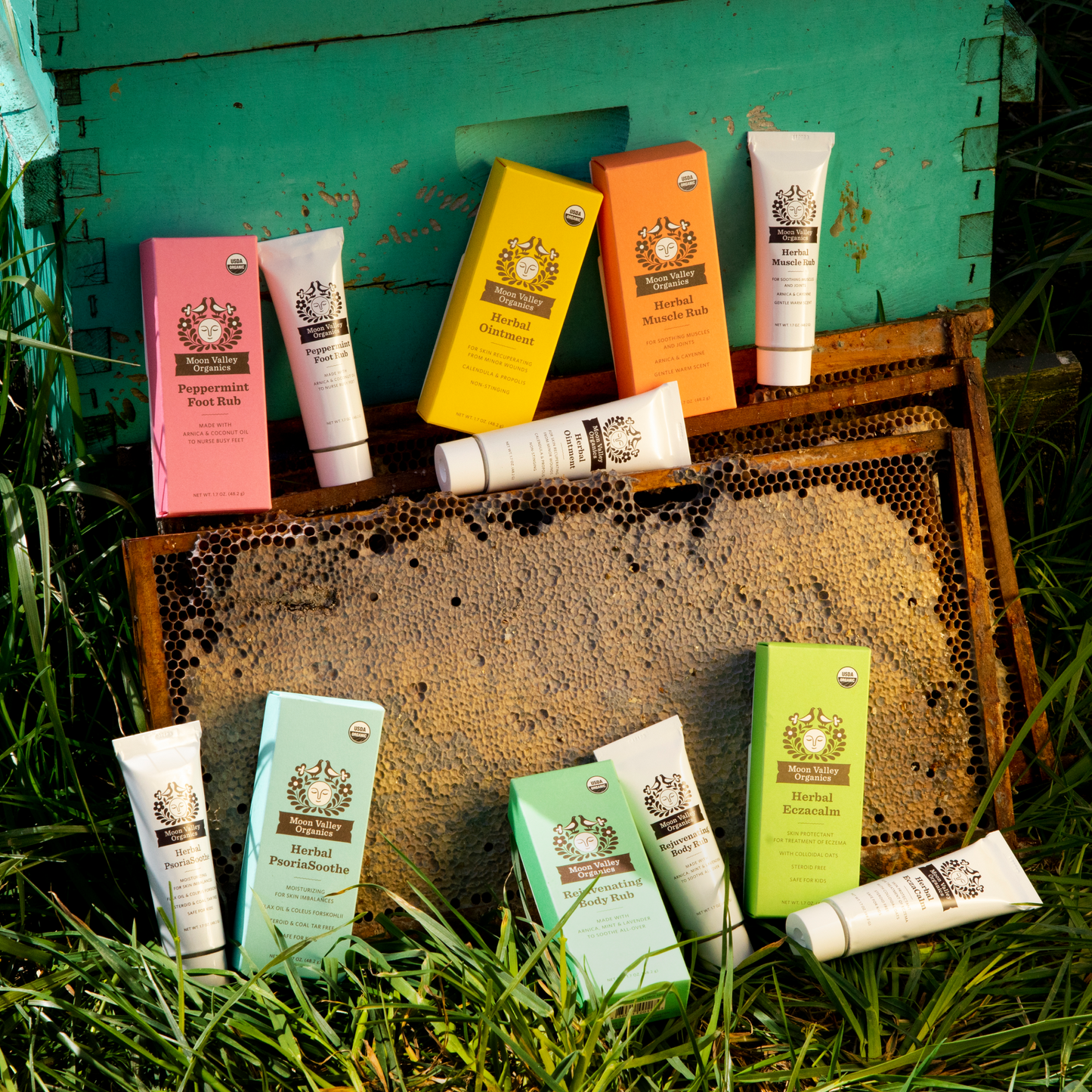 Moon Valley Organics Salves and Balms All boxes and tubes laying in the grass against a bee box and honeycomb